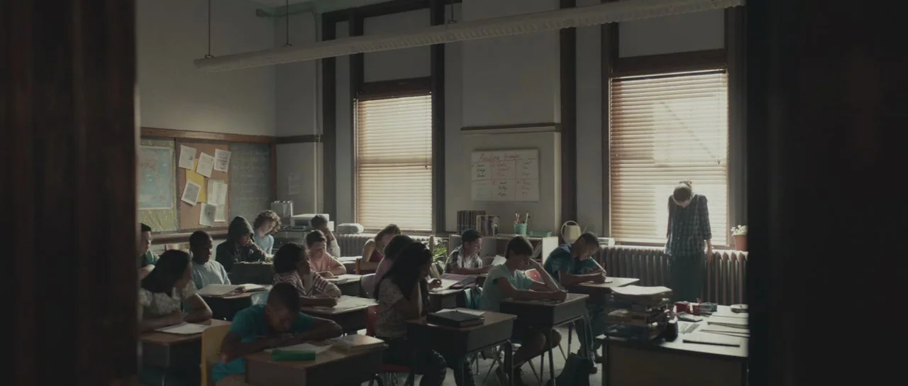 Meadowland (2015) movie still: wide — A traditional school classroom filled with rows of desks and seated elementary students. A teacher stands by…