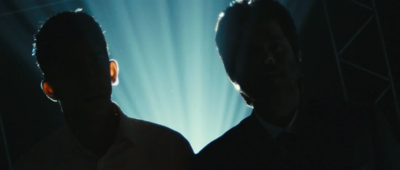 Slumdog Millionaire (2008) movie still: medium — A dark, cramped interior space is illuminated by a sharp beam of projector light. Two men are…