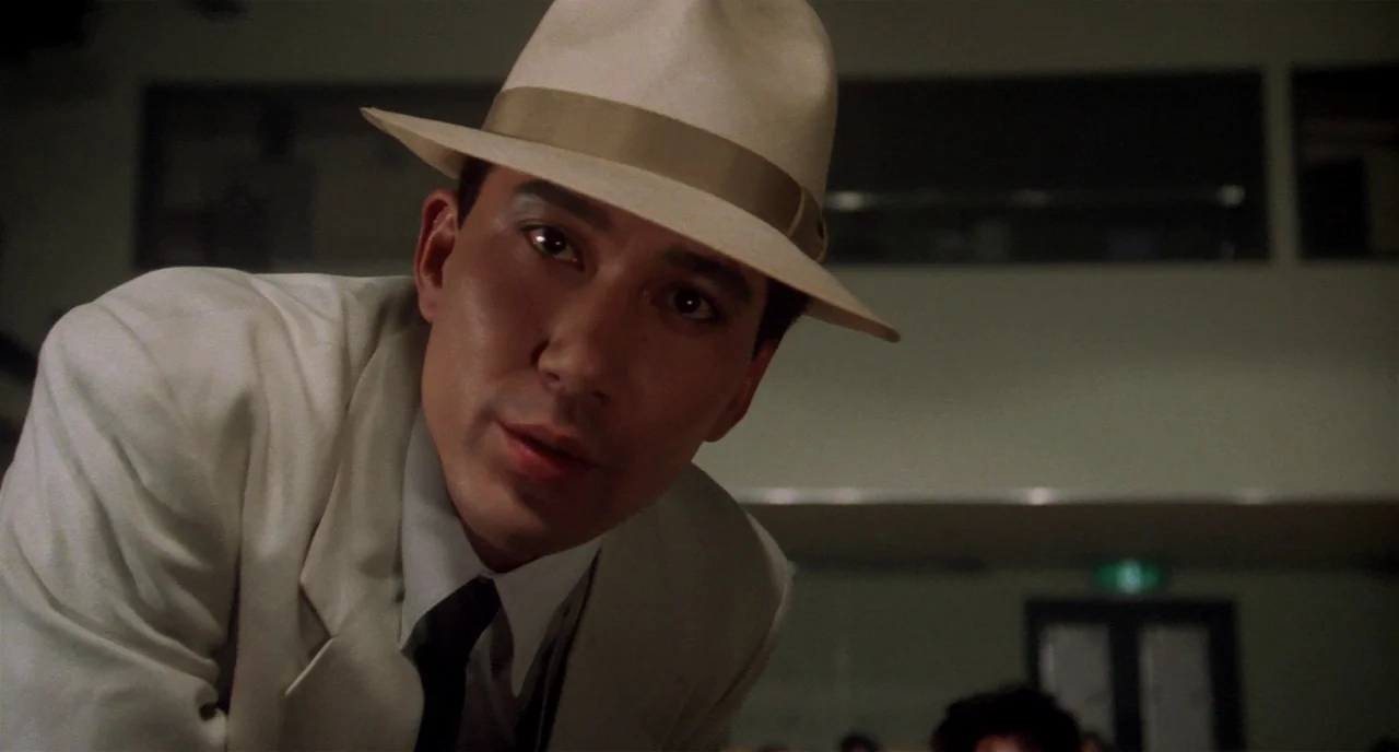 Tampopo (1985) movie still: closeup — An interior space with dim, out-of-focus background rows of seats in a large hall. A man wearing a white suit…