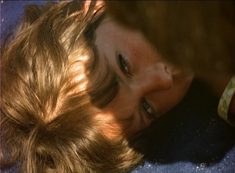 La Collectionneuse (1967) movie still: closeup — A person lies on a dark blue towel covered in scattered grains of sand. The subject rests their…