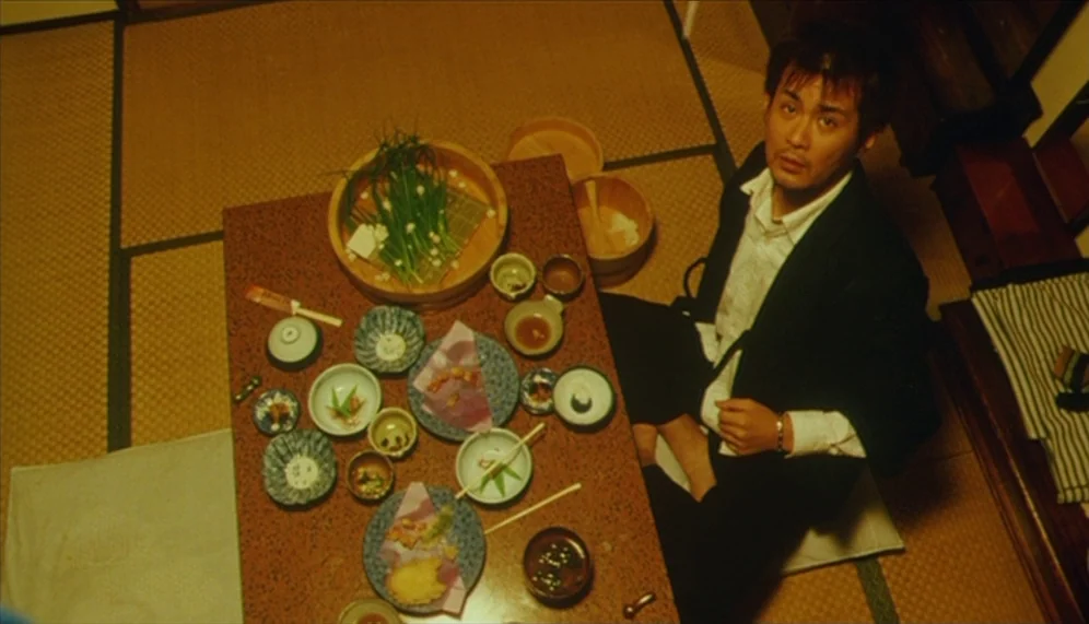 Gozu (2003) movie still: medium — A traditional Japanese room features tatami mat flooring and a low dining table. A man in a suit sits beside a…