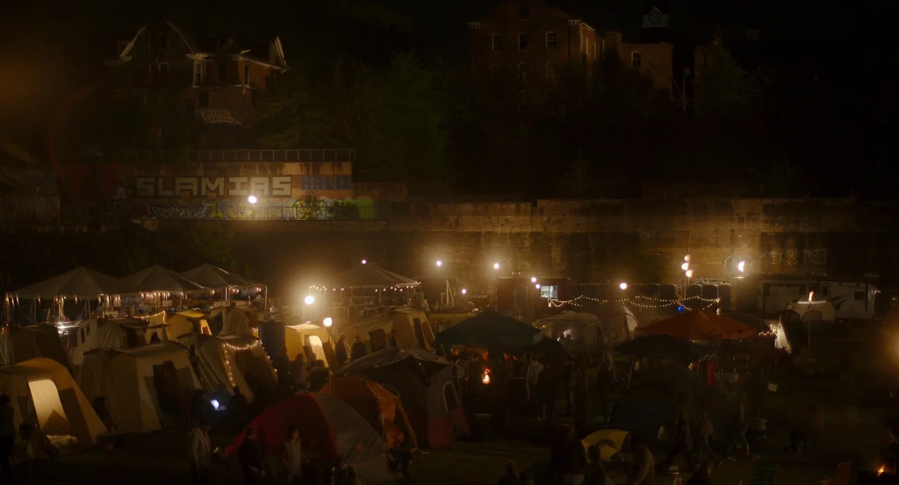 Civil War (2024) movie still: wide — A dense collection of camping tents occupies an open field beneath a high stone wall. The nighttime setting…