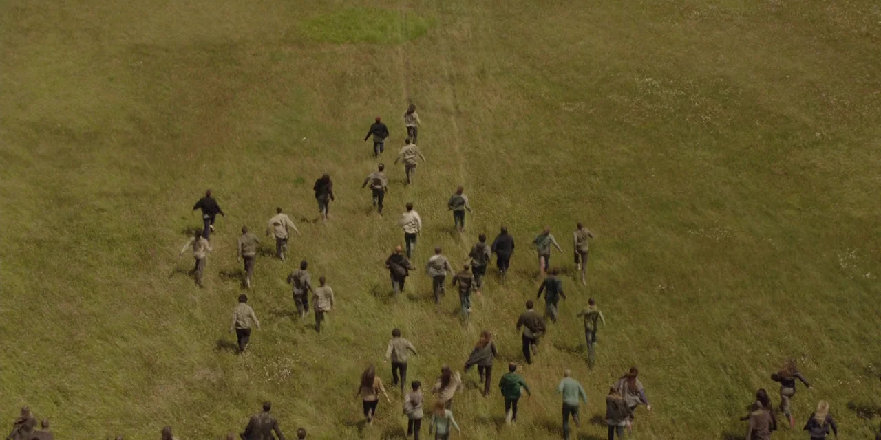 The Girl With All The Gifts (2016) movie still: wide — A vast, open meadow with long grass populated by a large group of people. Dozens of…