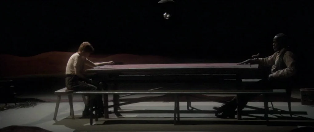 Manderlay (2005) movie still: wide — A long wooden table occupies a sparse and dimly lit interior space against darkness. Two men sit at the table…