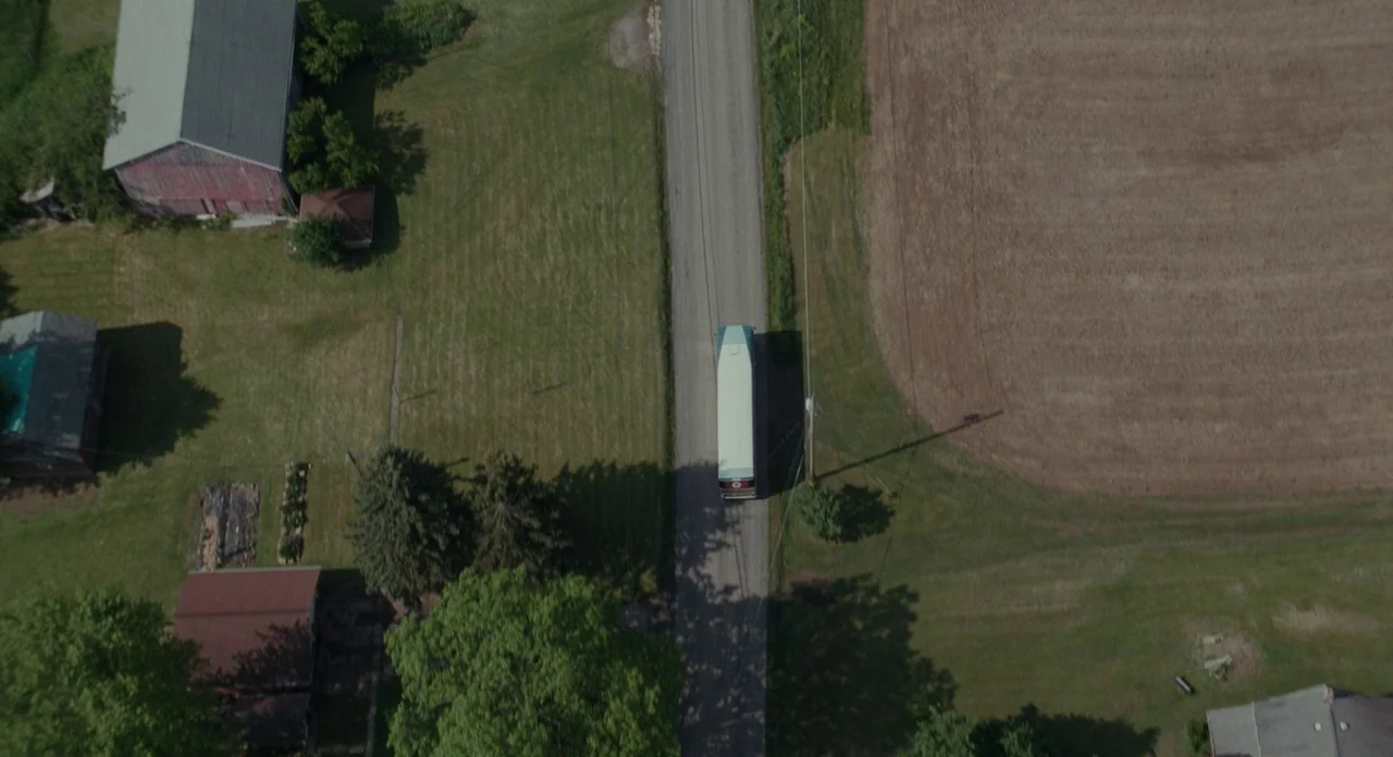 Promised Land (2012) movie still: wide — A top-down view shows a white bus traveling along a rural paved road. The road is bordered by a green grass…