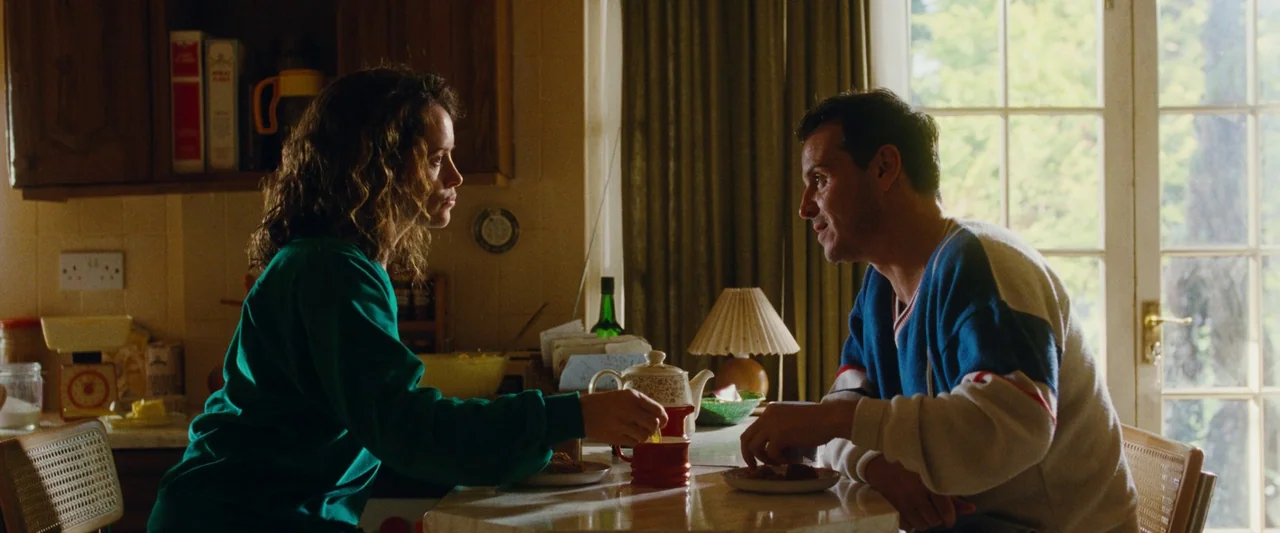 All Of Us Strangers (2023) movie still: medium — A sunlit indoor dining area with a white table and large window. A man and a woman sit facing each…