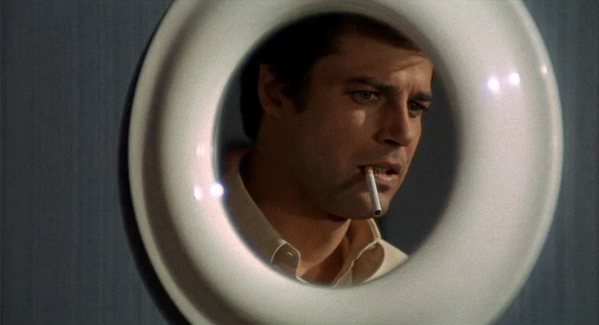 Perversion Story (1969) movie still: closeup — A man's face appears centered within the frame of a circular door peephole lens. He holds a lit…
