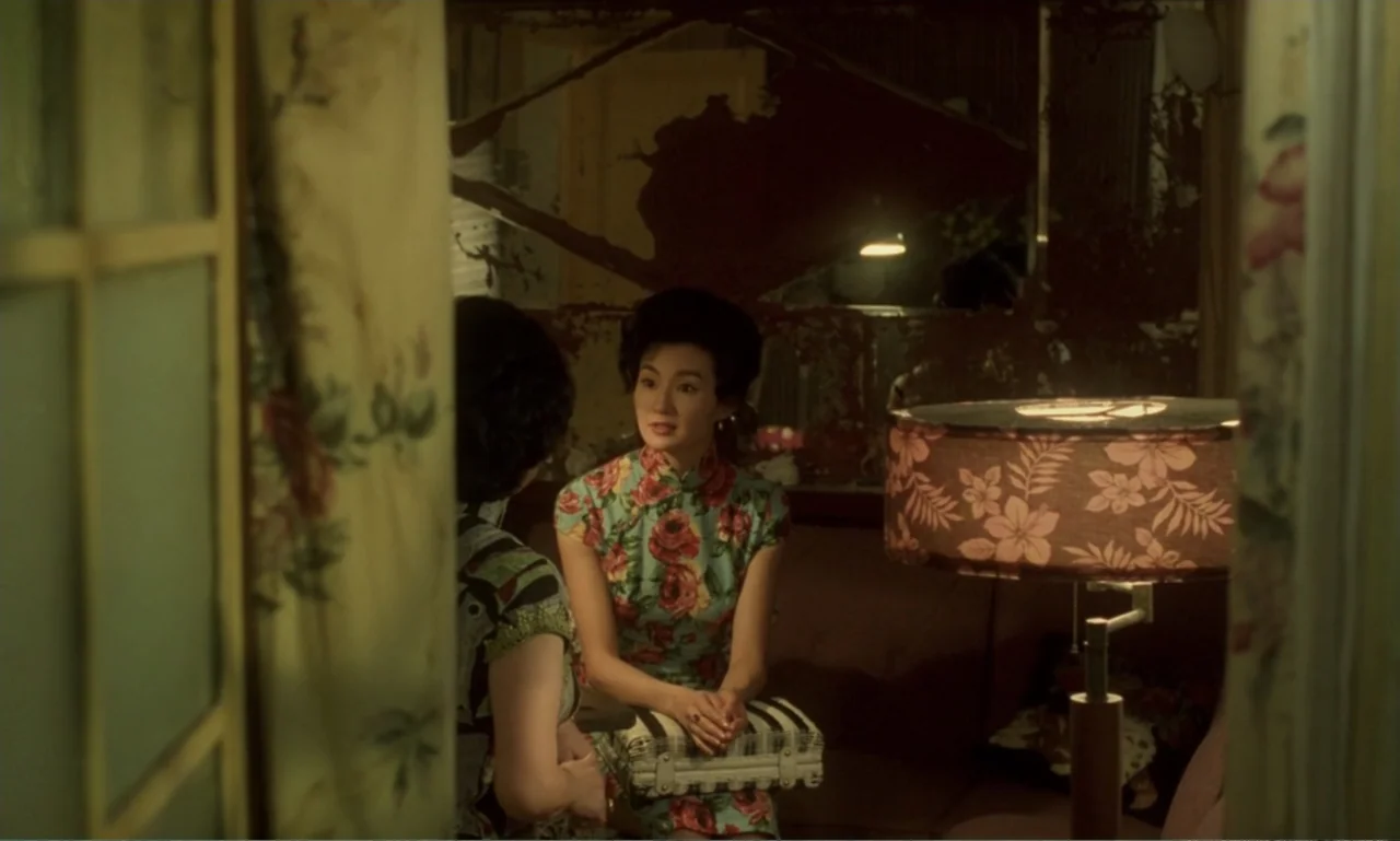 In the Mood for Love (2000) movie still: medium — A 1960s interior space features patterned fabric walls and a large, decorative table lamp. Two…