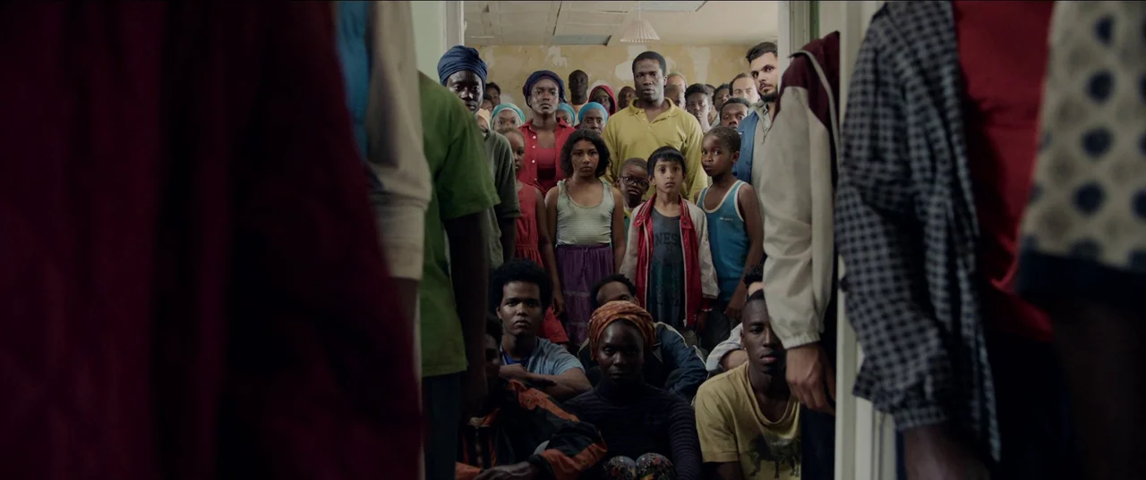 His House (2020) movie still: medium — A crowded residential interior space is filled with people of varying ages. The group stands and sits close…