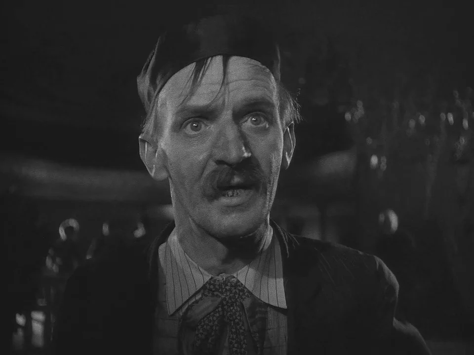 Sawdust and Tinsel (1953) movie still: closeup — A dimly lit room with a man in a beret as the focal point. The man stares ahead with a wide-eyed…