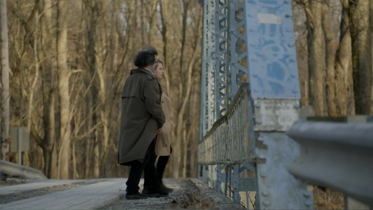 Murders at the Burger Joint (2022) movie still: fullbody — A man and a woman stand on a weathered metal bridge over a wooded area. The two…