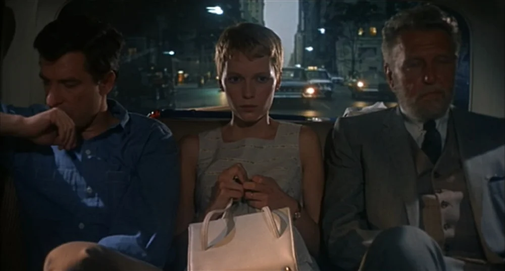 Rosemary’s Baby (1968) movie still: medium — Three individuals sit side by side in the back seat of a dark taxi. City streets are visible through…