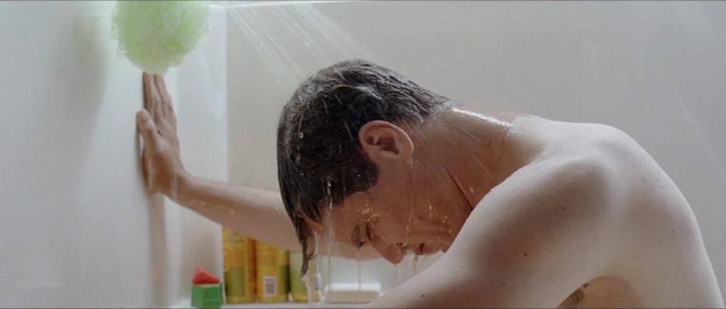 Take Shelter (2011) movie still: medium — A modern shower stall features white walls and a green mesh bath sponge. A man stands under the running…