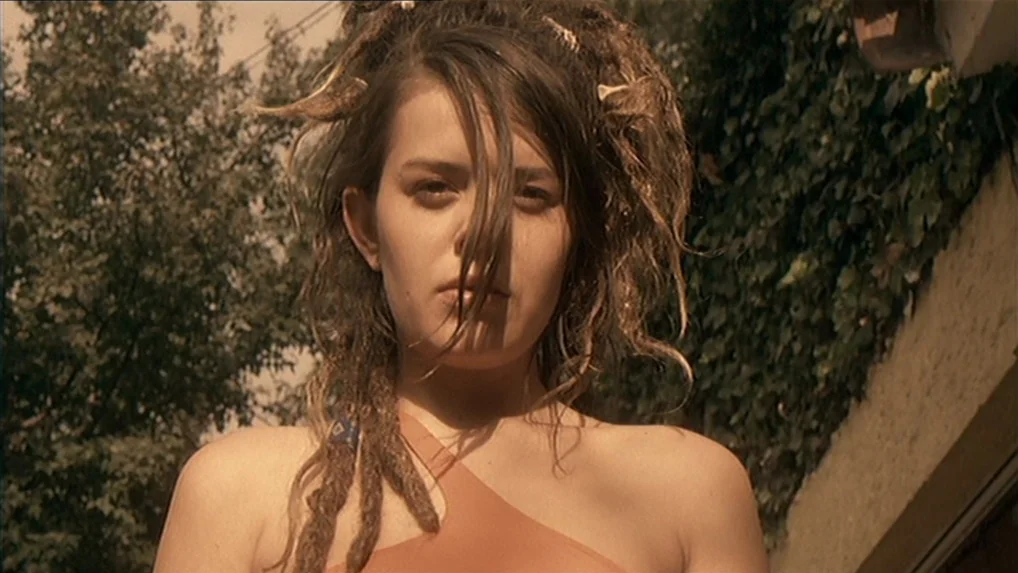 Battle In Heaven (2005) movie still: closeup — An outdoor setting features a vine-covered wall and tree foliage in the background. A young woman…