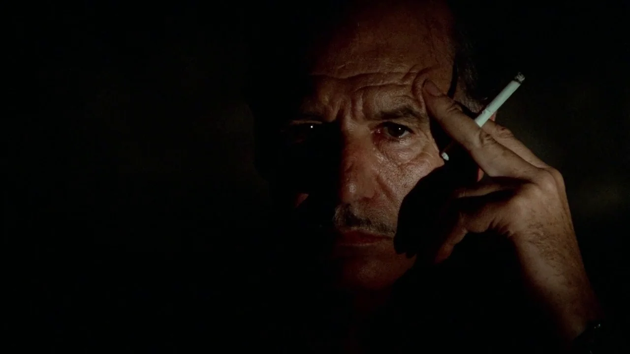 Sorcerer (1977) movie still: closeup — An elderly man is framed against a near-total black void in a dark room. He holds a lit cigarette to his…