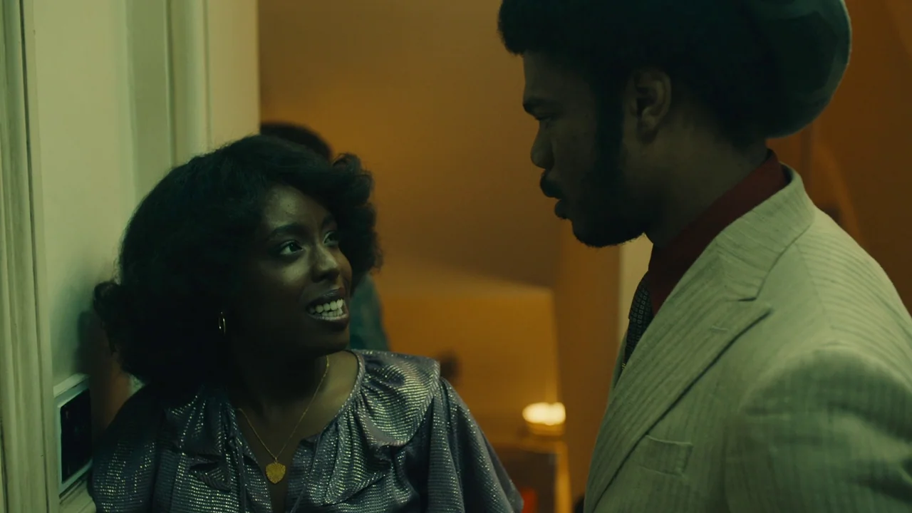 Lover’s Rock (2020) movie still: medium — An interior hallway with cream-colored walls and soft, dim lighting conditions. A woman facing the camera…