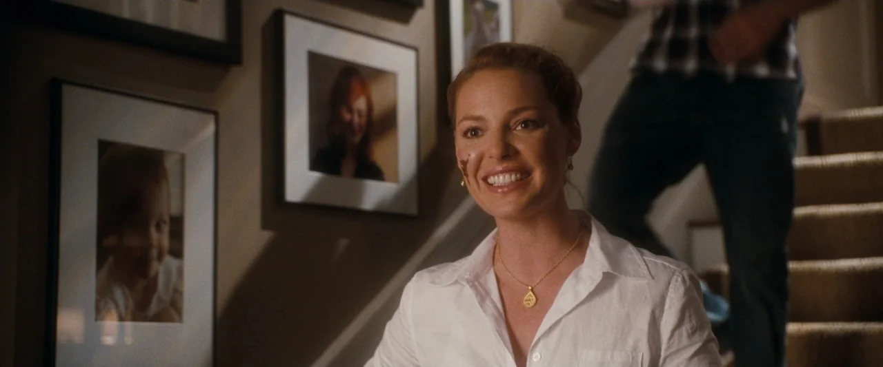 Life As We Know It (2010) movie still: medium — A residential staircase landing features framed photographs hung on a wall. A woman smiles broadly…