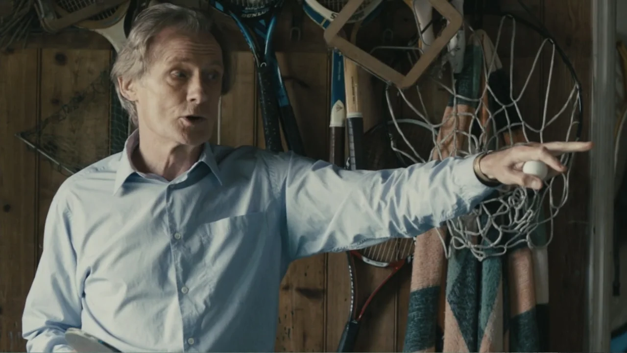 About Time (2013) movie still: medium — A garage interior featuring wooden walls cluttered with various hanging sports rackets and equipment. A man…