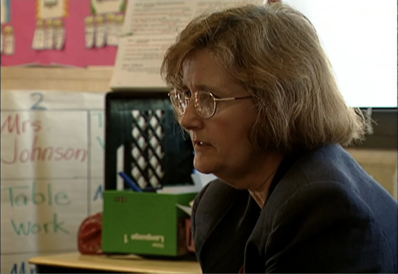 Broken Child (2000) movie still: medium — A school classroom features a whiteboard with handwritten instructional text in the background. A woman in…