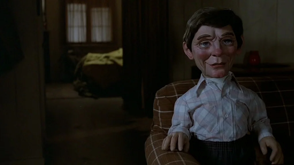 Magic (1978) movie still: medium — A dark, dimly lit room features wood-paneled walls and a fabric-covered chair. A wooden ventriloquist dummy sits…