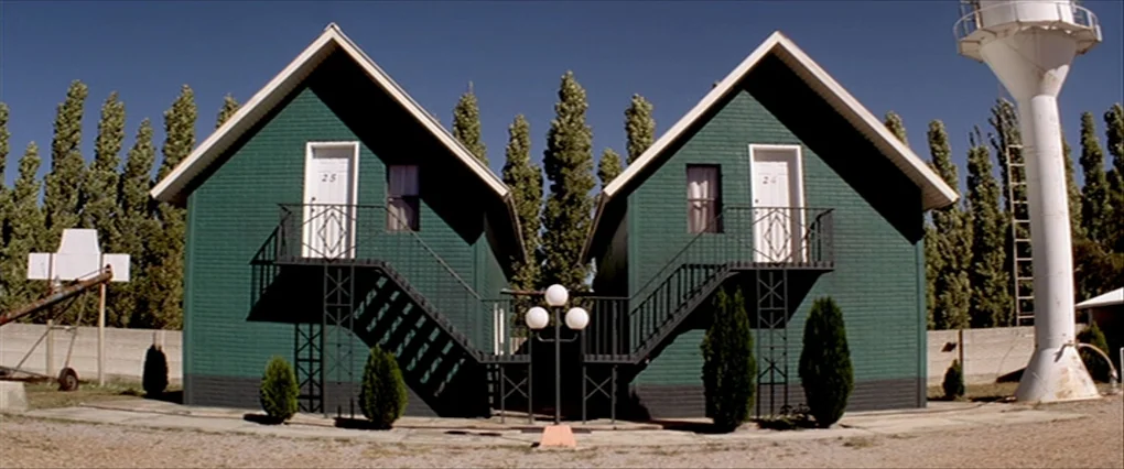 Silent Light (2007) movie still: establishing — Two identical green cabins with exterior staircases stand symmetrically in a gravel lot. A tall…