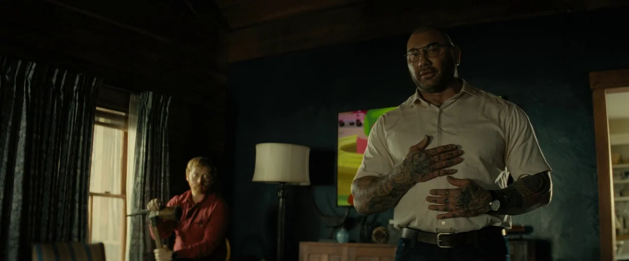 Knock at the Cabin (2023) movie still: medium — A rustic cabin with dark walls and a television screen visible in the background. One man stands…