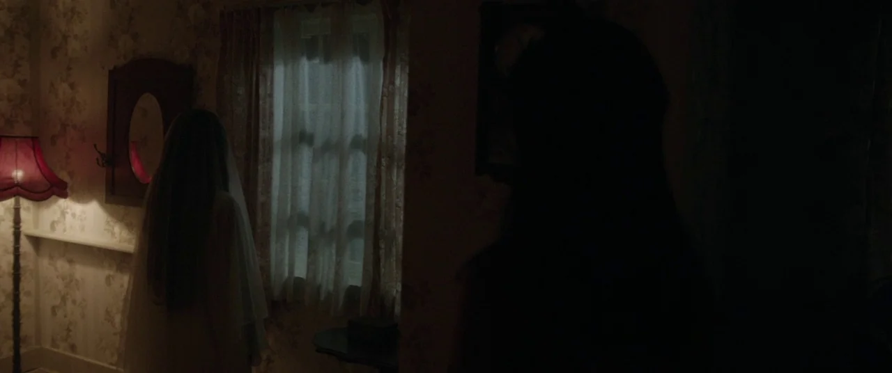 Satan’s Slaves (2017) movie still: medium — A dimly lit room features floral wallpaper and a window covered by white curtains. Two women stand in…