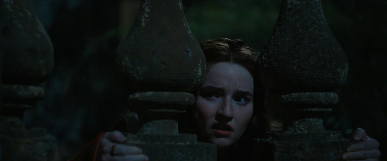 Rosaline (2022) movie still: closeup — A woman looks through a dark stone balustrade at night. Her face is partially obscured by the weathered stone…