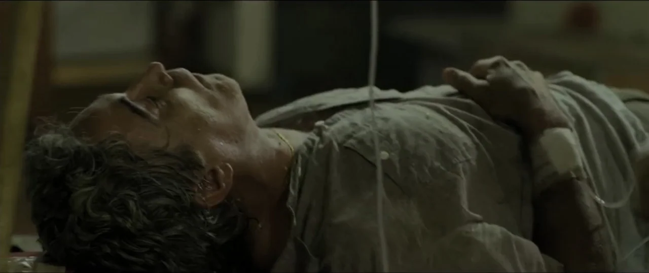 Primal (1994) movie still: medium — A dimly lit hospital room with a patient lying on a narrow bed. A middle-aged man with graying hair rests with…