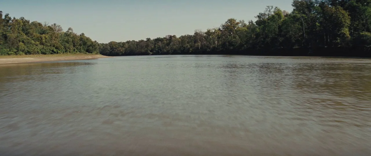 Mud (2012) movie still: establishing — A wide, muddy river flows through a dense forest under a clear sky. Thick tree lines border the water on both…
