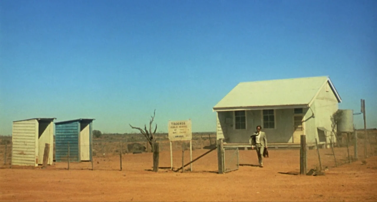 Wake In Fright (1971) movie still: wide — A remote, isolated schoolhouse stands in a vast, dry, red-dirt desert landscape. A man in a suit walks…