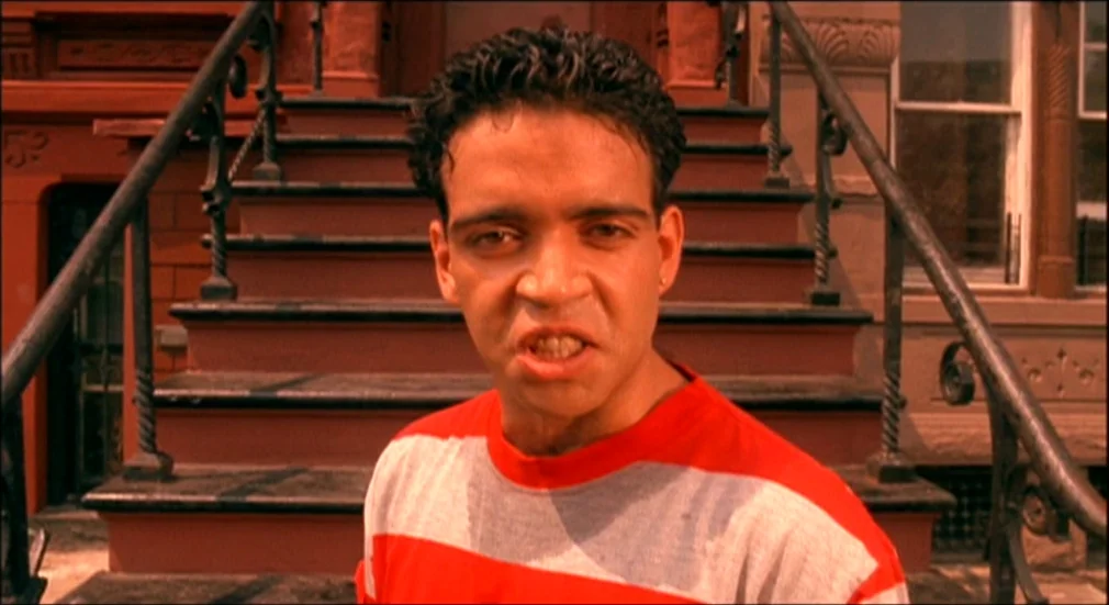 Do The Right Thing (1989) movie still: medium — A man stands in front of a red brick building with dark metal stair railings. He looks forward…