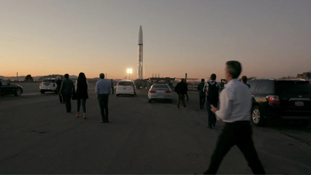 Wild Wild Space (2024) movie still: wide — A wide paved runway features several parked cars and a tall rocket launch vehicle.