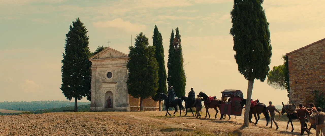 Benedetta (2021) movie still: wide — A rural Italian landscape features a stone church standing amidst tall cypress trees. A small group of men on…