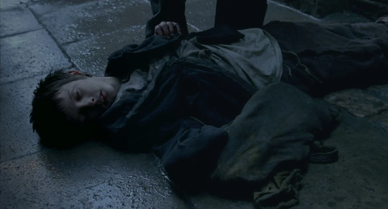 The Pianist (2002) movie still: fullbody — A wet, dark cobblestone ground serves as the backdrop for a prone child. An adult figure stands above the…