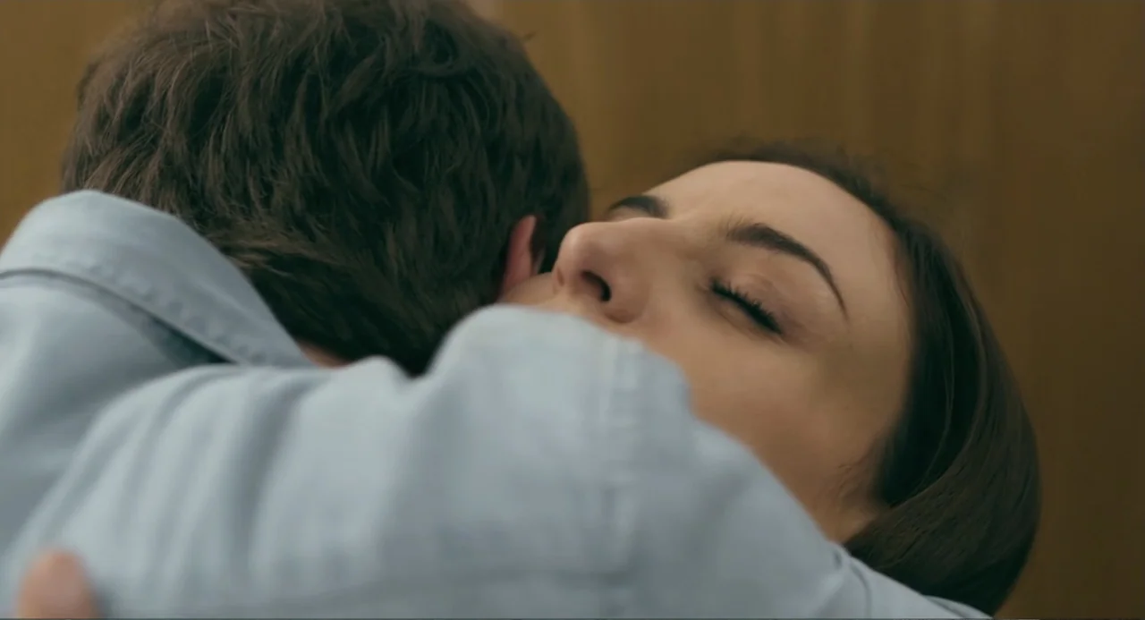The Lift (2021) movie still: closeup — A close-up shot focuses on a woman's face during an embrace inside a room. She holds a person whose back is…