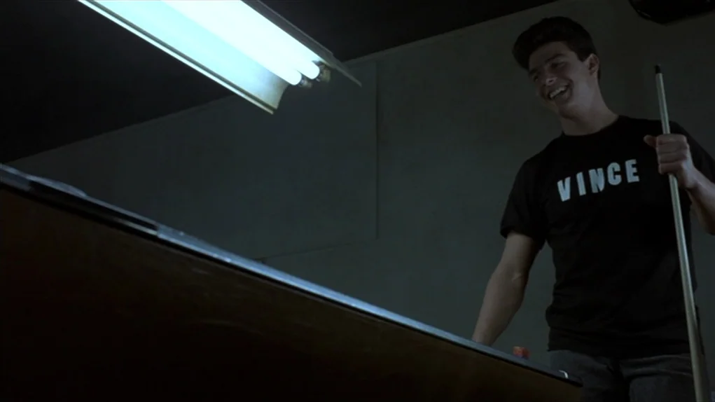 The Colour of Money (1986) movie still: medium — A dim room features a low-hanging fluorescent light over a pool table surface. A young man wearing…