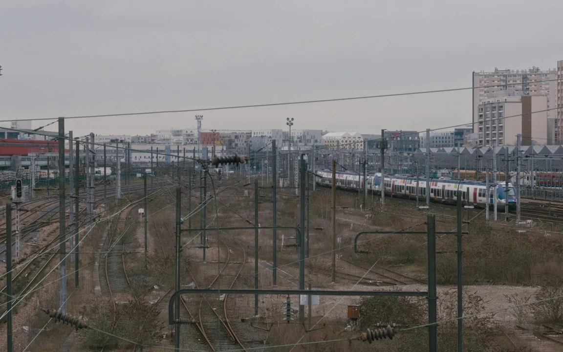 Let The Sunshine In (2017) movie still: establishing — An expansive industrial railway yard stretches toward a background of mid-rise urban…