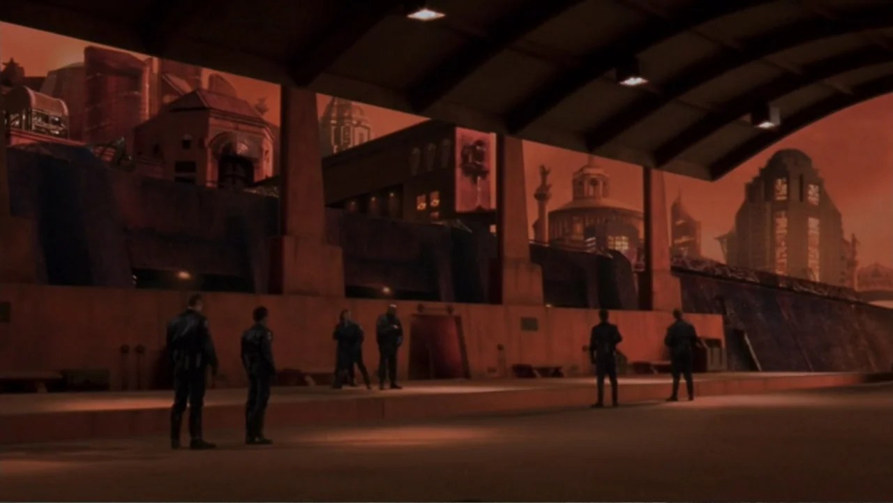 Ghosts of Mars (2001) movie still: wide — A wide shot of a futuristic transit platform at dusk with a sprawling cityscape. Six uniformed individuals…