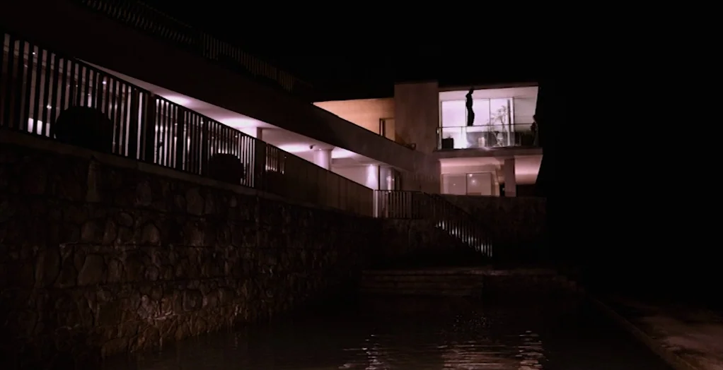 Dollhouse (2012) movie still: establishing — A modern multi-level villa with stone walls sits above a pool at night. The structure is illuminated…