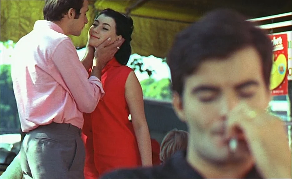 Le Bonheur (Happiness) (1965) movie still: medium — An outdoor cafe setting features a couple interacting in the background focus. A man with a pink…