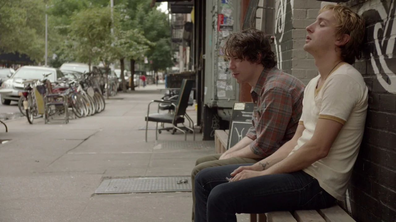 The Heart Machine (2014) movie still: medium — A city sidewalk beside a brick wall covered in layered black and white graffiti. Two men sit on a…