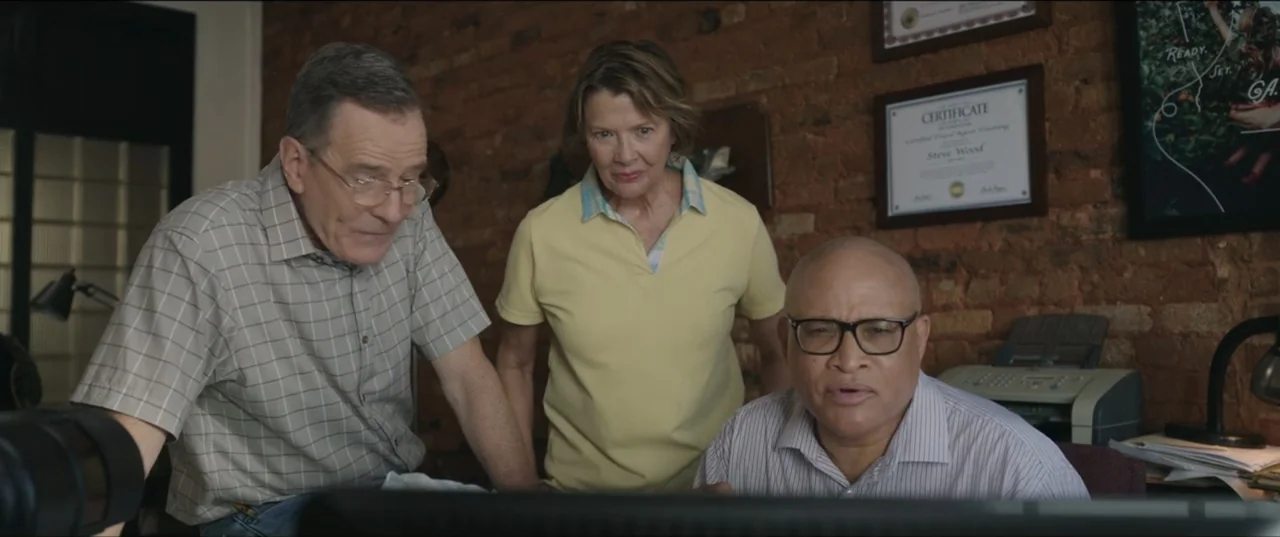 Jerry & Marge Go Large (2022) movie still: medium — An office features exposed brick walls and framed certificates hanging on the wall. Three people…