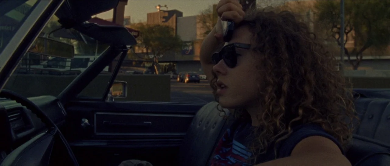 Bliss movie still: medium — A person sits in the driver seat of a convertible during golden hour. The background shows a commercial building parking…