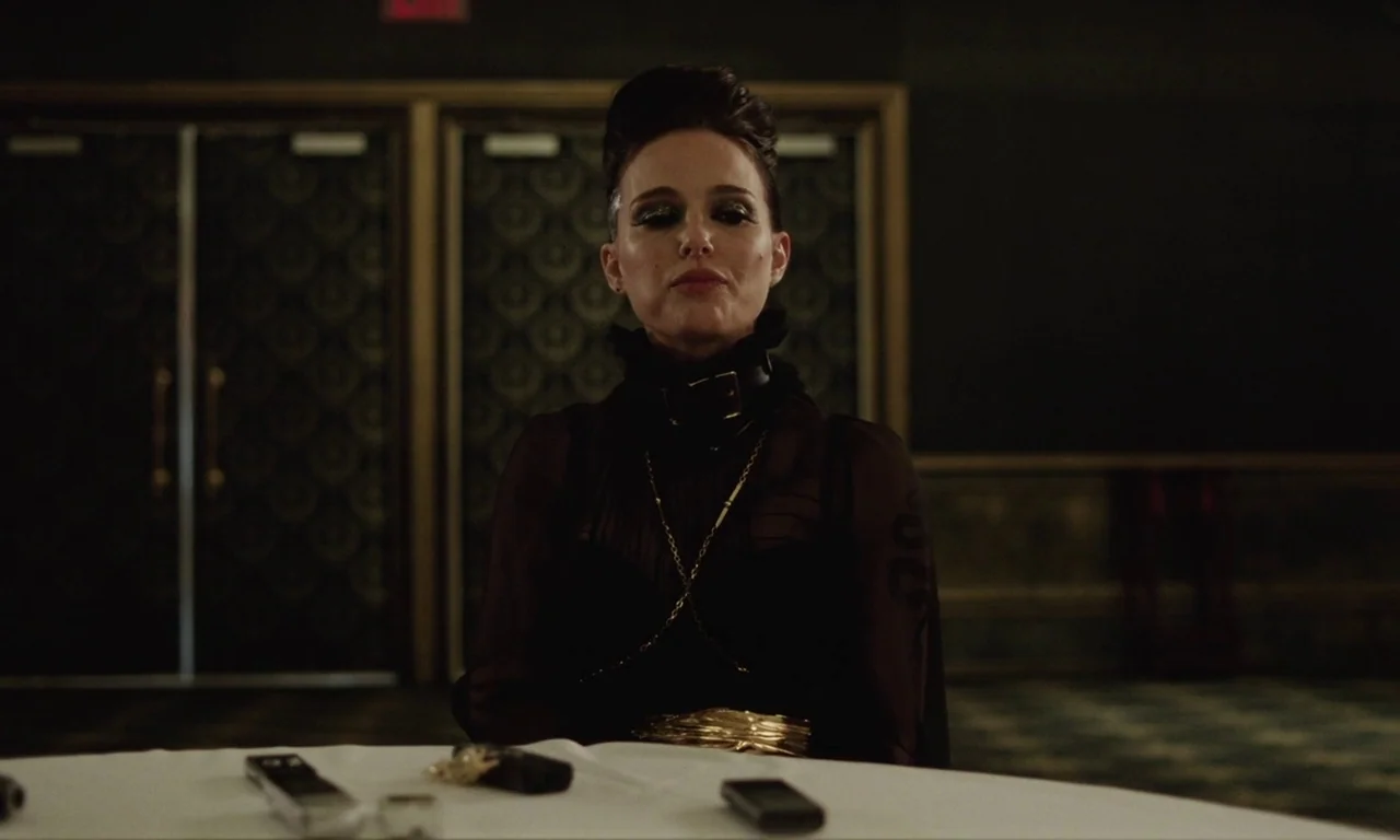 Vox Lux (2018) movie still: medium — A dimly lit hotel ballroom features patterned double doors in the background. A woman sits alone at a round…