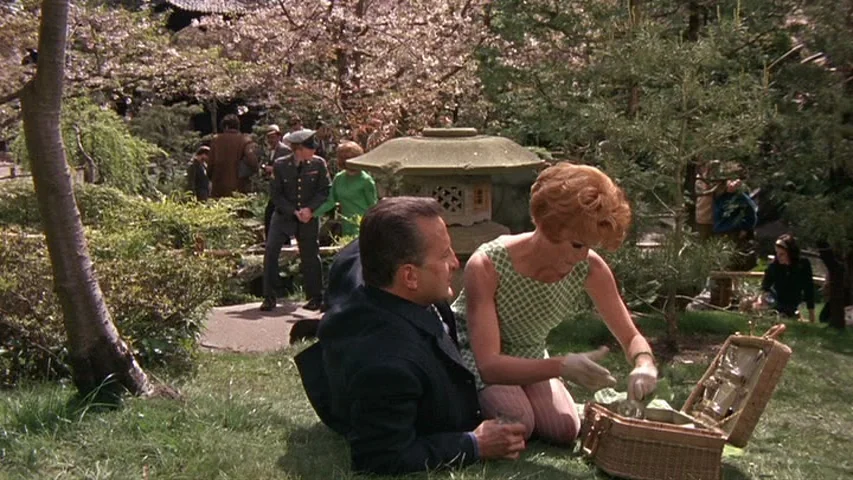 Petulia (1968) movie still: medium — A man and woman sit on the grass in a lush park setting. The woman interacts with an open picnic basket while…