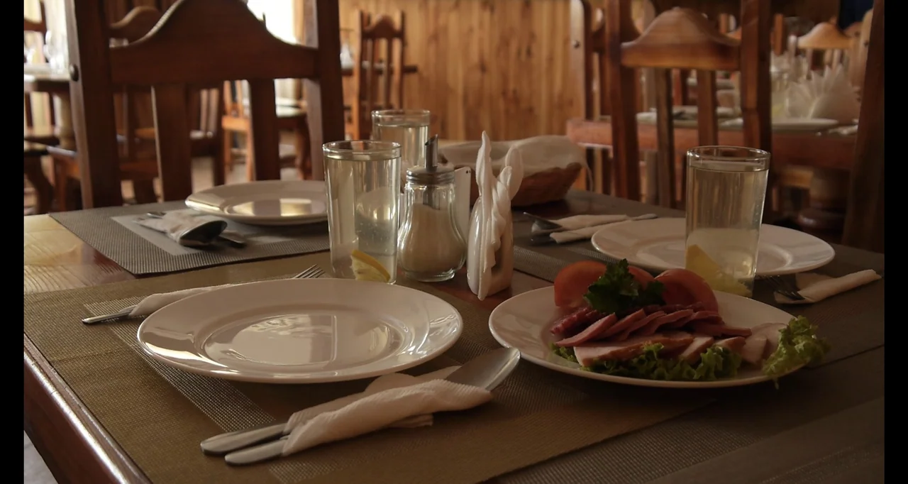 Chernobyl's Café (2016) movie still: medium — A wooden restaurant table is set with white plates, water glasses, and sliced meats. Several empty…