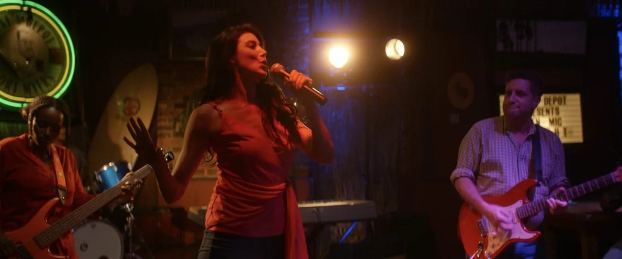 On a Wing and a Prayer (2023) movie still: medium — A dimly lit musical venue features a live band performing on a small stage. The singer stands…