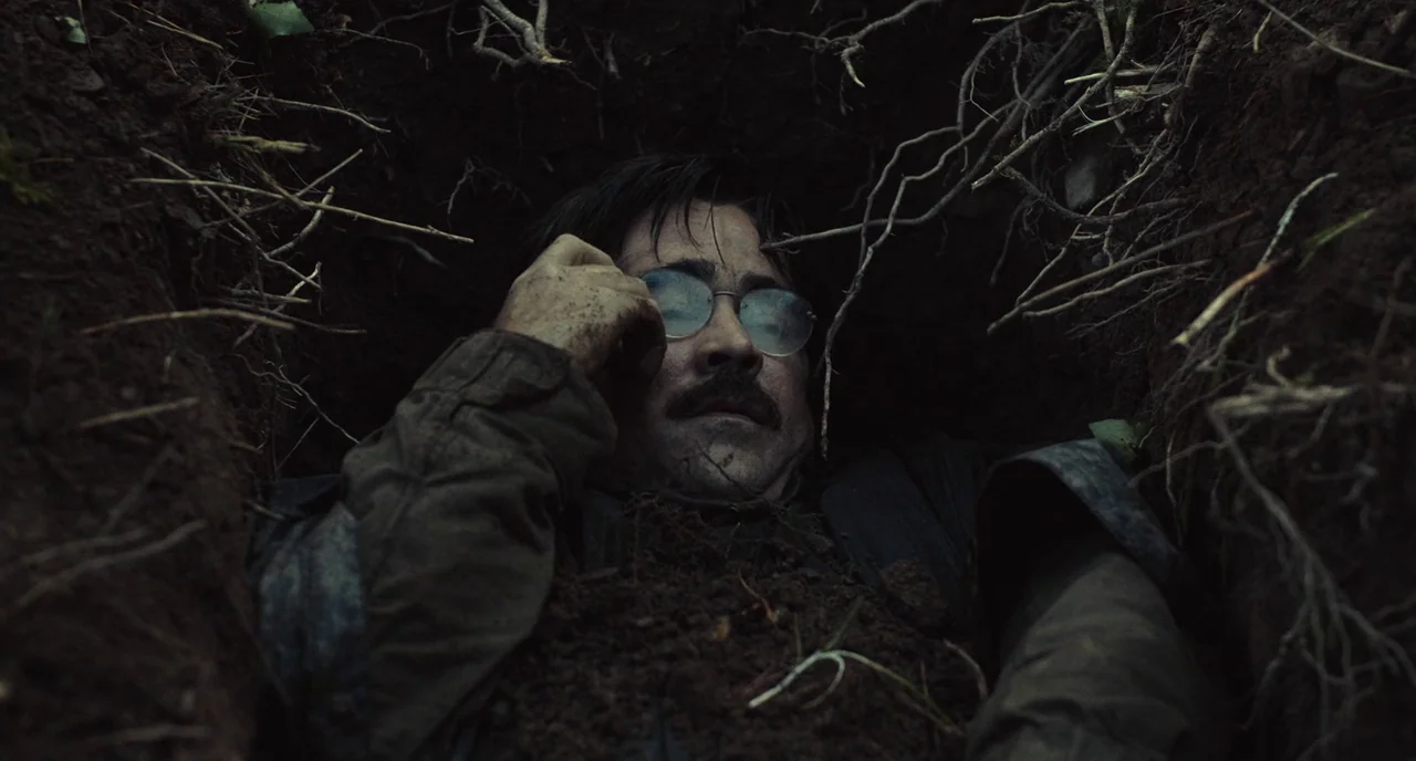 The Lobster (2015) movie still: closeup — A cramped, excavated hole in the earth lined with exposed roots and loose soil. A man in a military jacket…
