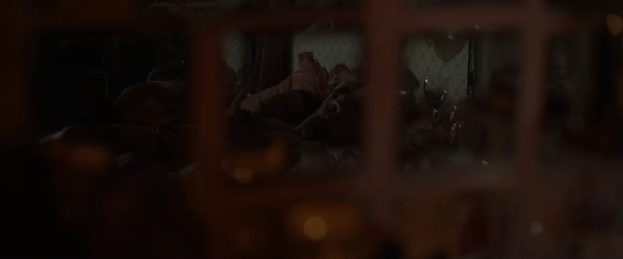 Small Crimes (2017) movie still: medium — A dark bedroom viewed through a glass window pane with light reflections. A man reclines in bed next to a…